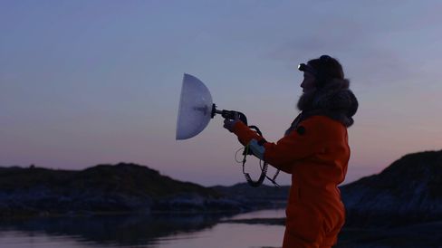 Person in orange jacket holding a device with a half-round reflective dish by a body of water at dusk