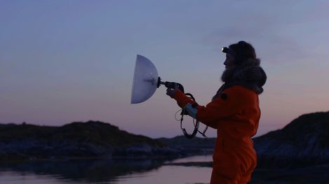 Person in orange jacket holding a device with a half-round reflective dish by a body of water at dusk