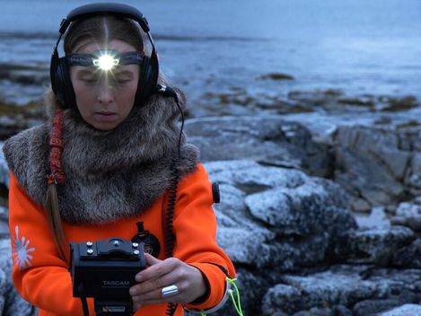 Person wearing headphones and a fur cloak holding a recording device in front of rocky coastline