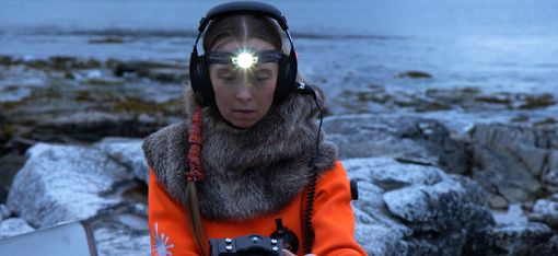 Person wearing headphones and a fur cloak holding a recording device in front of rocky coastline