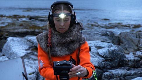 Person wearing headphones and a fur cloak holding a recording device in front of rocky coastline