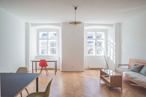 Bright room with parquet floor, two large windows, a table with a red chair, a sofa, and a coffee table