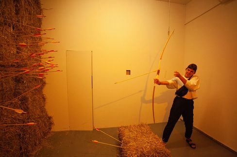 Person in a room with hay bales aiming a bow at a wall covered with many arrows.