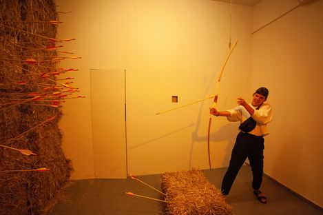 Person in a room with hay bales aiming a bow at a wall covered with many arrows.