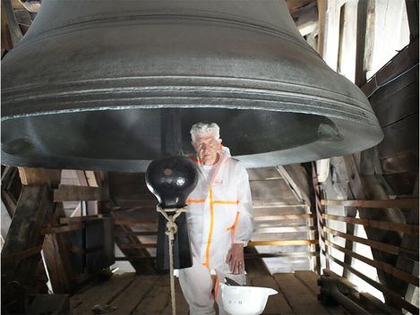 Person in weißem Schutzanzug steht unter einer großen Glocke in einem Holzturm.