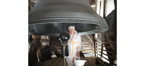 Person in weißem Schutzanzug steht unter einer großen Glocke in einem Holzturm.