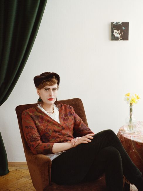Person sitting with folded hands in a brown armchair next to a table with a vase of yellow flowers against a white wall with a small picture