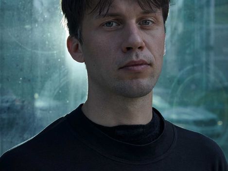 Portrait of a young man with dark hair wearing a black sweater in front of a glass facade in an urban setting.