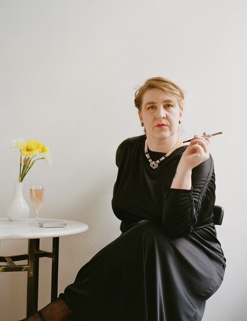 Man in black dress sitting with crossed legs on a chair holding a cigarette in her right hand next to a small white table with a vase of yellow flowers and a glass.