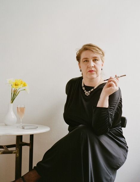 Man in black dress sitting with crossed legs on a chair holding a cigarette in her right hand next to a small white table with a vase of yellow flowers and a glass.
