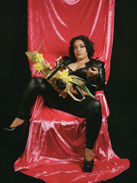 Person in black leather clothing sitting on a chair draped with red fabric holding a chainsaw decorated with yellow flowers
