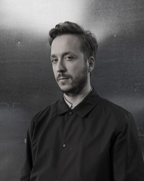 Portrait of a man with short hair and beard against a dark wall.