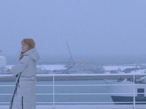 Person mit Mantel lehnt an Reling eines Schiffs mit Blick auf Hafen und Wasser im Hintergrund