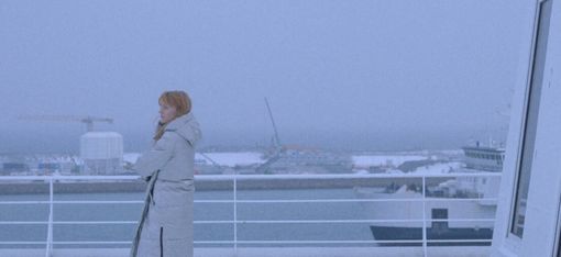Person mit Mantel lehnt an Reling eines Schiffs mit Blick auf Hafen und Wasser im Hintergrund