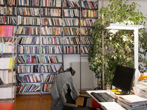 Arbeitszimmer mit vollregaligem Bücherwand, Schreibtisch mit Computer und Bürostuhl mit Jacke.