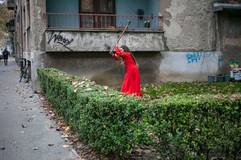Woman in red dress hitting a hedge with a hammer in front of an old apartment building on a city street.