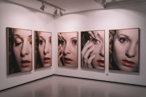 Five large portrait photographs of a woman applying makeup in detail displayed on a white gallery wall.