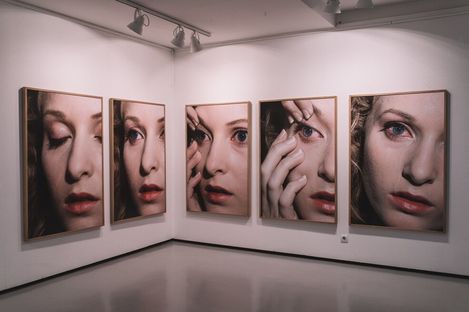 Five large portrait photographs of a woman applying makeup in detail displayed on a white gallery wall.