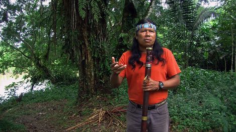 Man with long hair wearing an orange T-shirt stands in a forest holding a long carved wooden staff