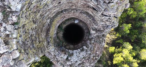 Blick von oben in eine tiefe, runde, mit Steinen gemauerte Turmruine umgeben von Bäumen