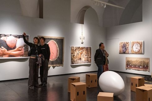 People in an exhibition room with several large artworks on the walls and a large white egg on the floor surrounded by simple cube seats.