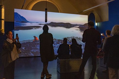 People sitting and standing in front of a large screen showing a landscape of water and mountains inside a room with an arched ceiling