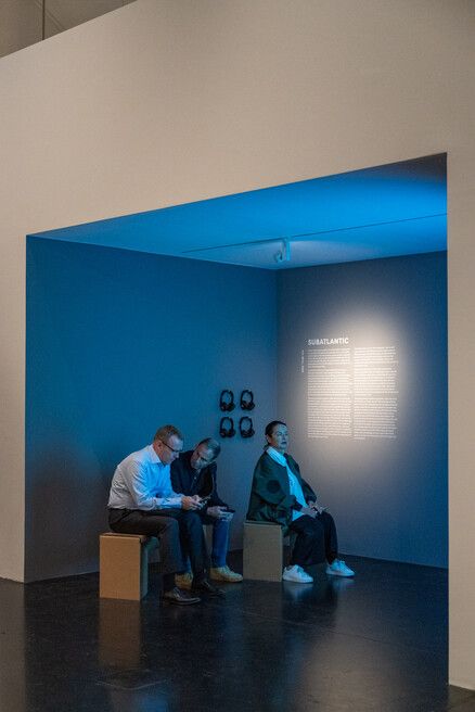 Three people sit on simple stools in a dark room lit with blue light, facing a wall illuminated with text and four mounted headphones.