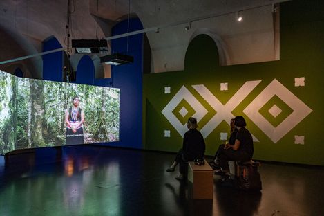Three people sit on benches in a dark room watching a large screen showing a video of a person in a forest, with a large white geometric pattern on a green wall.