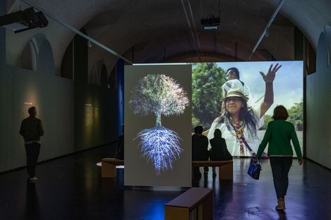 In a dark exhibition room, a lit projection of a tree with visible roots stands in front of a large screen showing two people, with several people sitting and walking nearby.