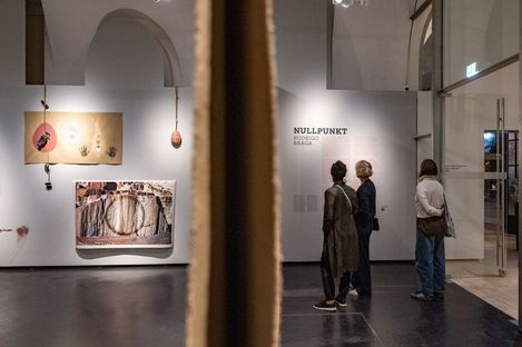 Three people looking at artworks and texts on a gallery wall with a vertical partition in the foreground