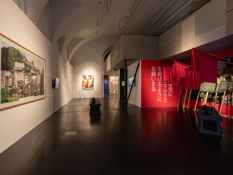 Art gallery with a modern exhibit; features a large photo, two sculptures, and vibrant red banners with Japanese text, creating a dynamic and thoughtful atmosphere.