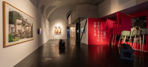 Art gallery with a modern exhibit; features a large photo, two sculptures, and vibrant red banners with Japanese text, creating a dynamic and thoughtful atmosphere.