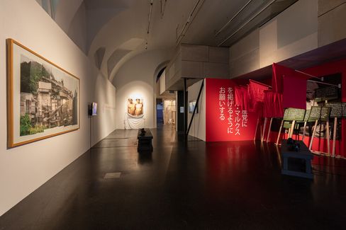 Art gallery with a modern exhibit; features a large photo, two sculptures, and vibrant red banners with Japanese text, creating a dynamic and thoughtful atmosphere.