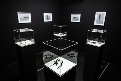 Five glass display cases with dark abstract objects on white pedestals in a room with several framed pictures on the walls.