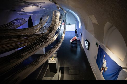 An art gallery with sweeping, black and white fabric installations hanging from the ceiling. A person observes exhibits along a curved wall, creating a contemplative atmosphere.