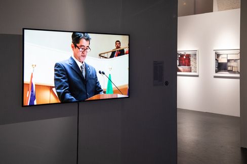 Flat screen displaying a man in a suit speaking at a podium with flags in the background in an art gallery with framed photos on white walls.