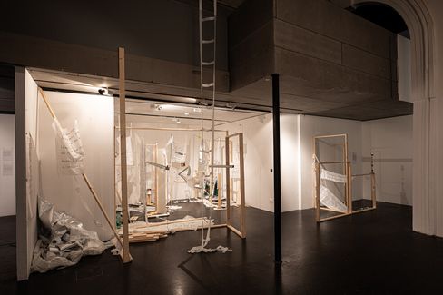 Installation view with multiple wooden frames, hanging fabric strips and a ladder in an exhibition space