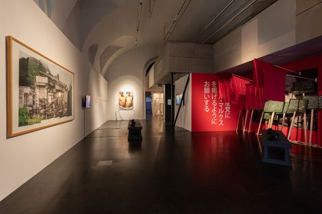 Art gallery with a modern exhibit; features a large photo, two sculptures, and vibrant red banners with Japanese text, creating a dynamic and thoughtful atmosphere.