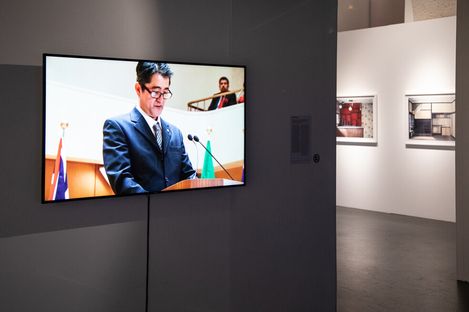Flat screen displaying a man in a suit speaking at a podium with flags in the background in an art gallery with framed photos on white walls.