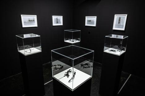 Five glass display cases with dark abstract objects on white pedestals in a room with several framed pictures on the walls.