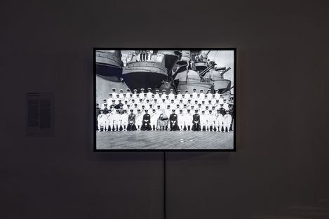 A black-and-white photo of a group of sailors in uniform, posed in rows on a ship deck. The backdrop features large ship structures, giving a historic, formal feel.
