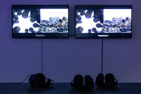 Dual screens display images of people peering downwards and an urban landscape in ruins. Headphones lie beneath, suggesting an interactive exhibit.