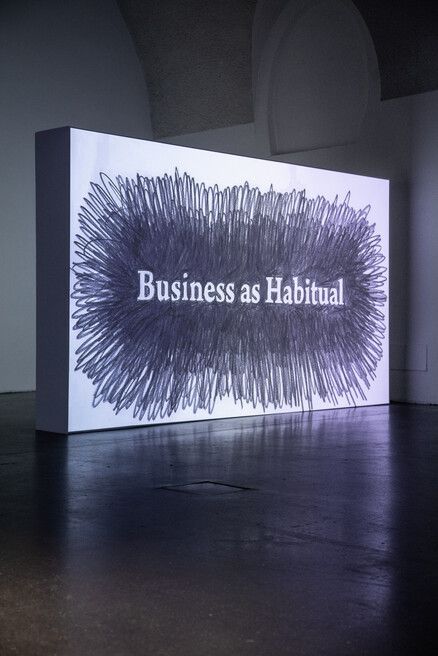 A thickly scribbled black text box with "Business as Habitual" in white letters is illuminated in a dimly lit room, conveying a tone of critique or reflection.