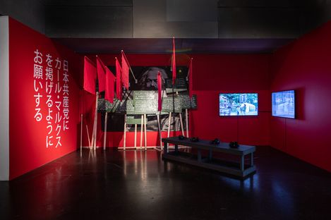 Exhibit with red walls features Japanese text, red flags, black-and-white signs, two screens displaying videos, and a bench with headphones. Mood: immersive.