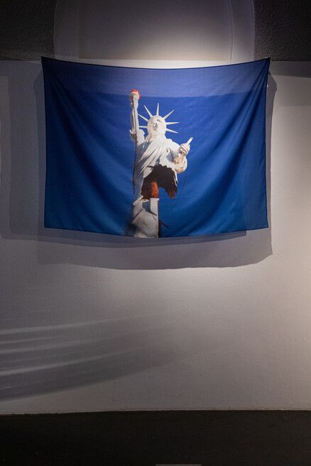 A tapestry hangs on a wall, depicting the Statue of Liberty against a deep blue background. The statue appears with its iconic torch and tablet.