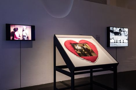 Gallery with two small screens showing videos on either side of a heart-shaped cushion display. The setting is modern and softly lit.
