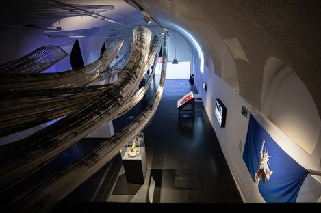 An art gallery with sweeping, black and white fabric installations hanging from the ceiling. A person observes exhibits along a curved wall, creating a contemplative atmosphere.