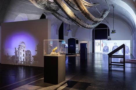 A dimly lit museum exhibit showcases artwork suspended from the ceiling, a glass display case, and a historical building projection, creating a contemplative atmosphere.