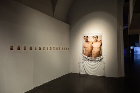 Two unclothed people with obscured faces in a large photograph hanging on a white wall, partially covered by a white cloth, with several smaller images arranged in a row beside it.