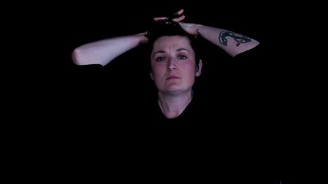 Woman with hair tied up and visible anchor tattoo on left upper arm against black background.